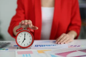 Pessoa de camisa vermelha aponta para um relógio de mesa vermelho em uma mesa com gráficos coloridos, simbolizando gestão de tempo e produtividade.