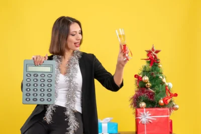 Mulher de terno comemorando resultados de campanhas de final de ano, segurando uma calculadora e uma taça de champagne ao lado de uma árvore de Natal decorada, representando a medição do ROI de campanhas de fim de ano.